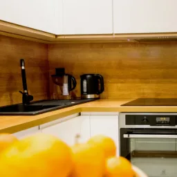 Nowoczesna kuchnia z białymi szafkami, drewnianym blatem i czarnym zlewem oraz czajnikiem elektrycznym, oświetlona ciepłym światłem.