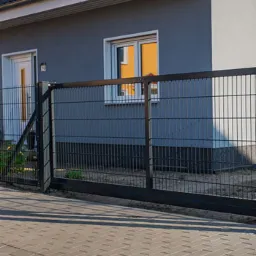 Nowoczesna, przesuwna brama wjazdowa z czarnego metalu, widoczna od strony posesji, z fragmentem ogrodzenia z siatki i szarą elewacją domu w tle.