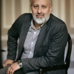 Portret mężczyzny z brodą, siedzącego na krześle w szarej marynarce i koszuli w paski, z zegarkiem na nadgarstku.