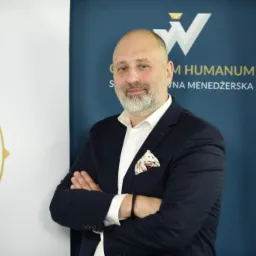 Portret mężczyzny w garniturze z poszetką w kieszonce, stojącego na tle banerów z logo Collegium Humanum.