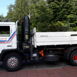 Biała ciężarówka MAN z dźwigiem HDS, zaparkowana obok domu z czerwonym dachem i zielonymi drzewami w tle.