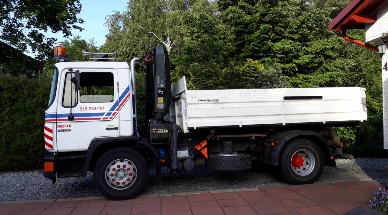 Biała ciężarówka MAN z dźwigiem HDS, zaparkowana obok domu z czerwonym dachem i zielonymi drzewami w tle.