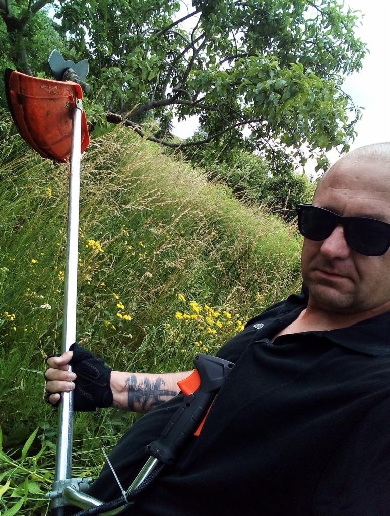 Mężczyzna w okularach przeciwsłonecznych pozuje z podkaszarką spalinową na tle wysokiej trawy i drzew