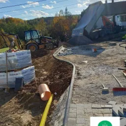 Przygotowanie terenu pod budowę chodnika z kostki brukowej: wykop z drenażem, krawężnik, maszyny budowlane i ciężarówka z kruszywem w tle.