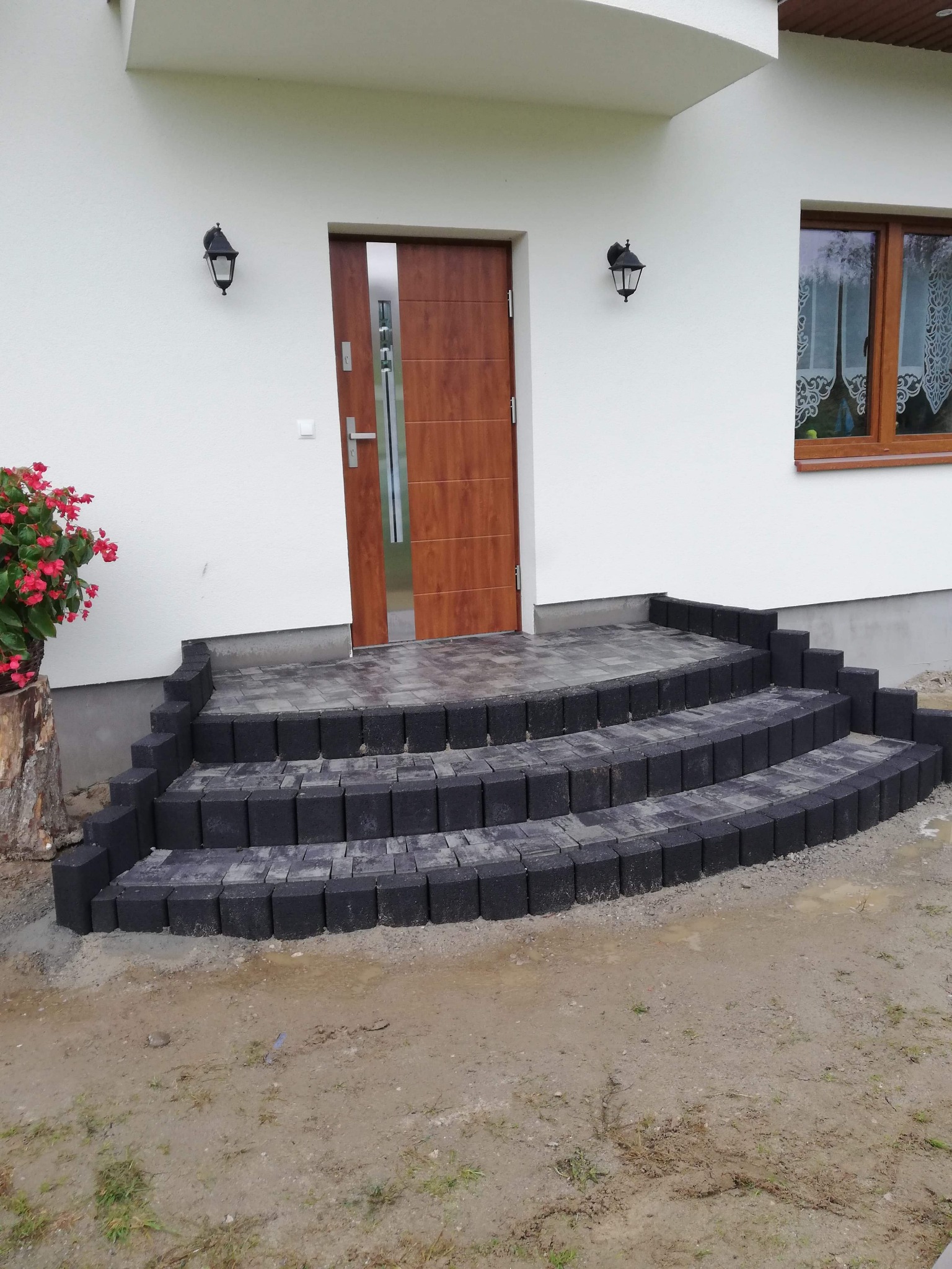 Schody wejściowe do domu wyłożone ciemnoszarą kostką brukową, z widocznym fragmentem drzwi wejściowych i kwiatami w donicy na pniu drzewa po lewej stronie, ziemia wokół schodów jeszcze nie...