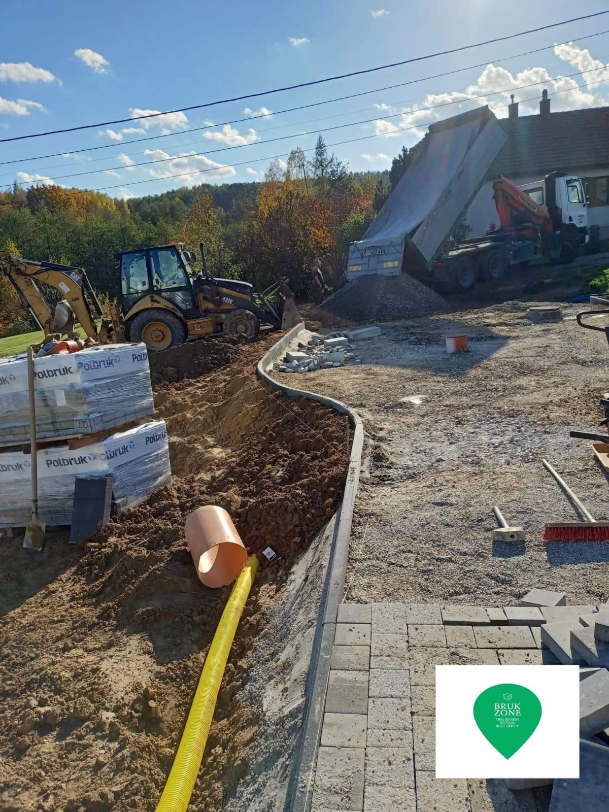 Przygotowanie terenu pod budowę chodnika z kostki brukowej: wykop z drenażem, krawężnik, maszyny budowlane i ciężarówka z kruszywem w tle.