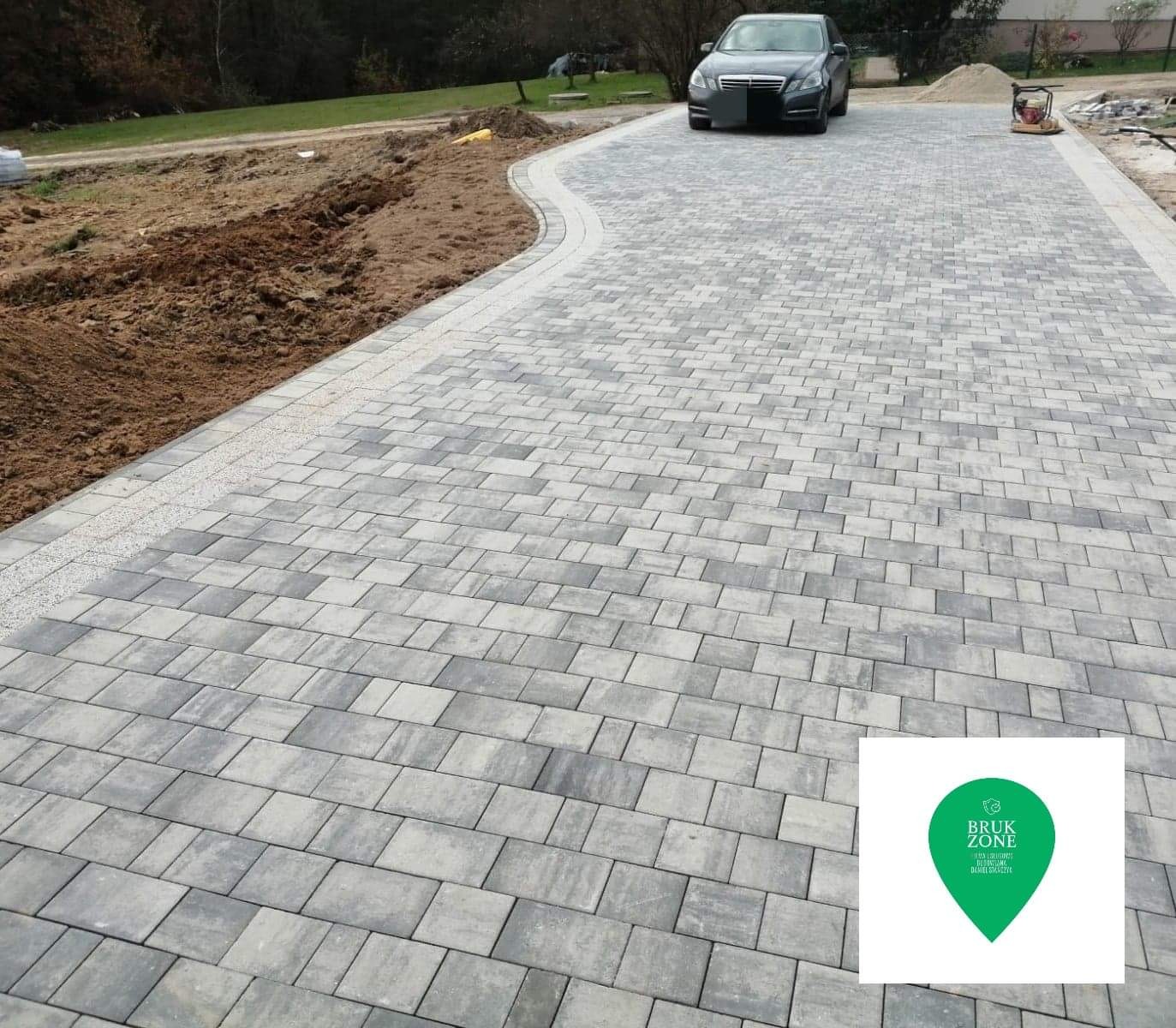 Ułożona szara kostka brukowa na podjeździe, z widocznym samochodem osobowym w tle i przygotowanym terenem pod dalsze prace brukarskie po lewej stronie.