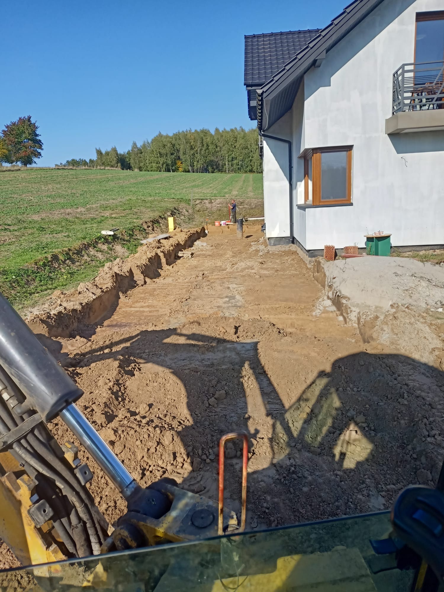 Wykop pod fundamenty domu jednorodzinnego, widok z kabiny koparki, wykopane rowy, przygotowany teren, pracownik w oddali.