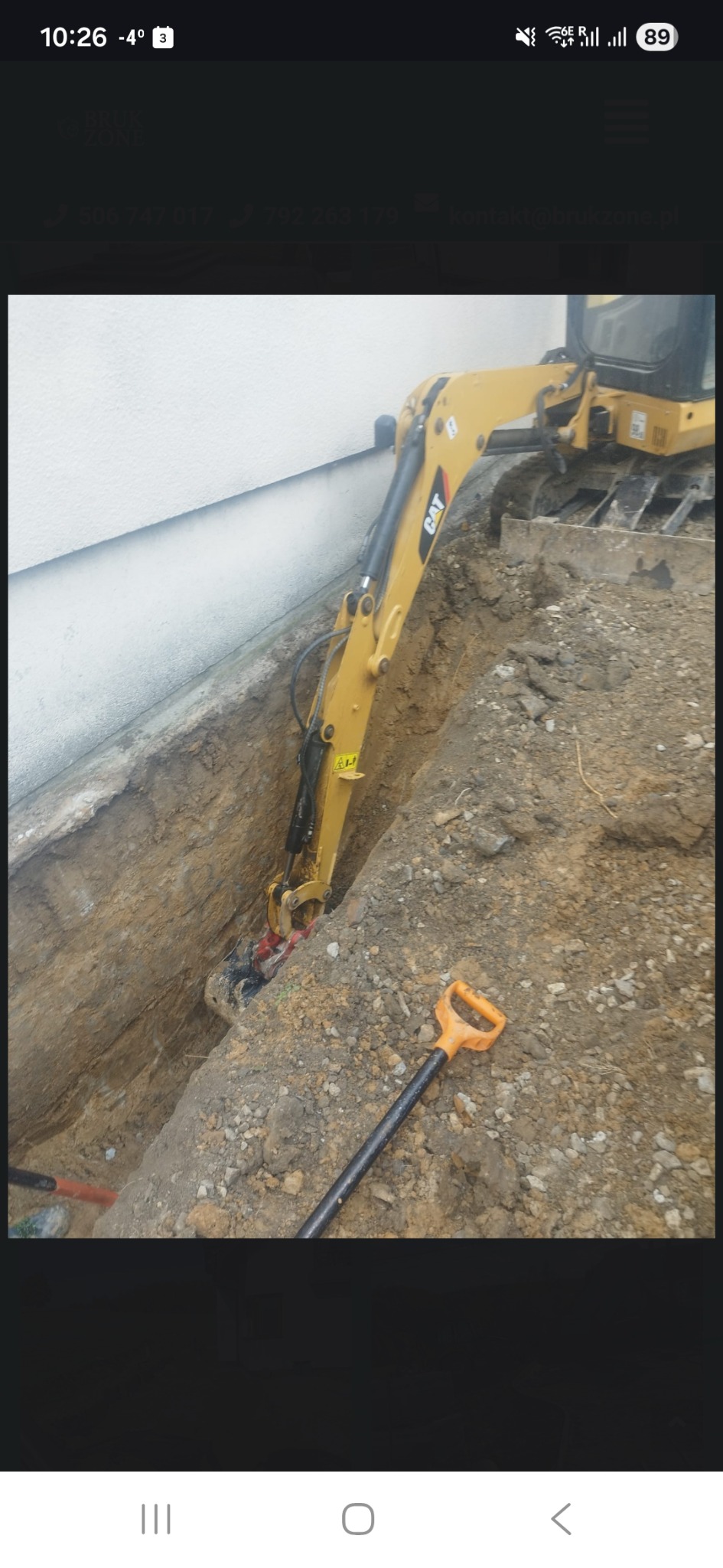 Żółta koparka CAT w wąskim wykopie przy ścianie budynku. Widoczna łopata i fragment rury. Prace ziemne, prawdopodobnie drenaż lub instalacje.