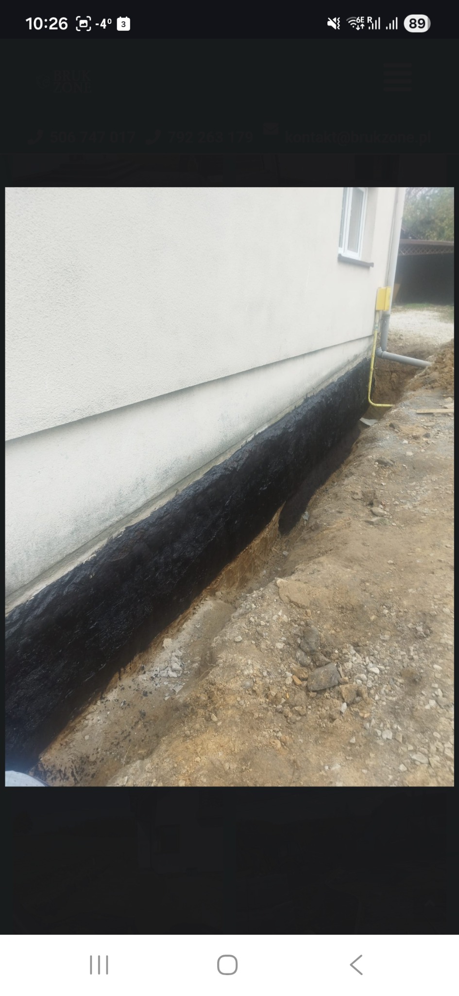 Zabezpieczona, czarną izolacją fundamentowa ściana budynku, widoczna w wykopie. Żółta rura gazowa przy ścianie. Grunt wokół budynku.