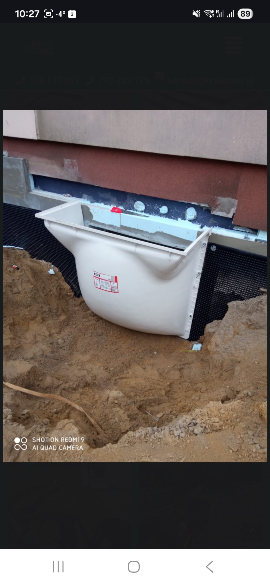 Montaż okna piwnicznego w wykopie z izolacją przeciwwilgociową. Widoczna czarna folia kubełkowa i ocieplenie fundamentu. Praca w toku, ziemia wokół.