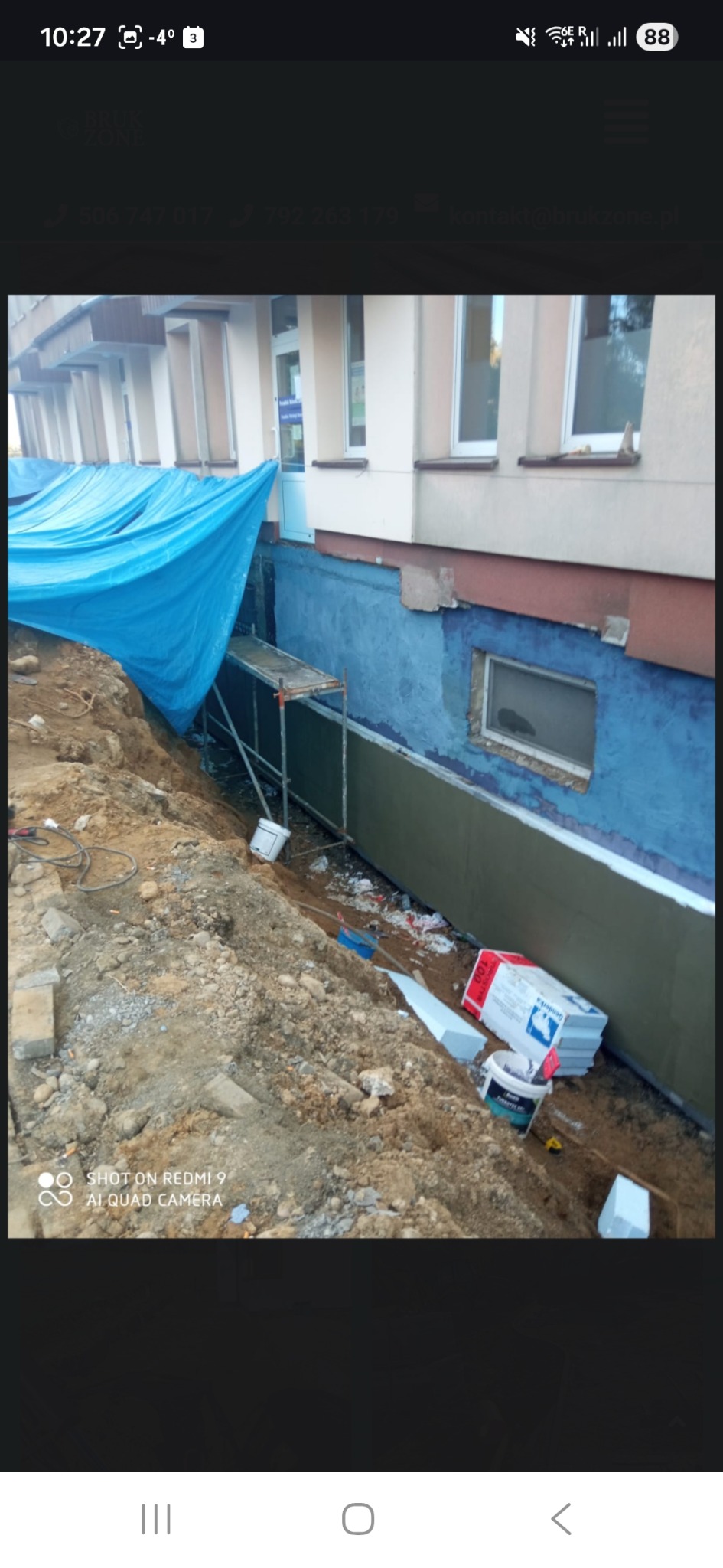 Odkopane fundamenty budynku z widocznym ociepleniem, rusztowaniem i plandeką. Częściowo pomalowana ściana na niebiesko. Widoczne materiały budowlane.