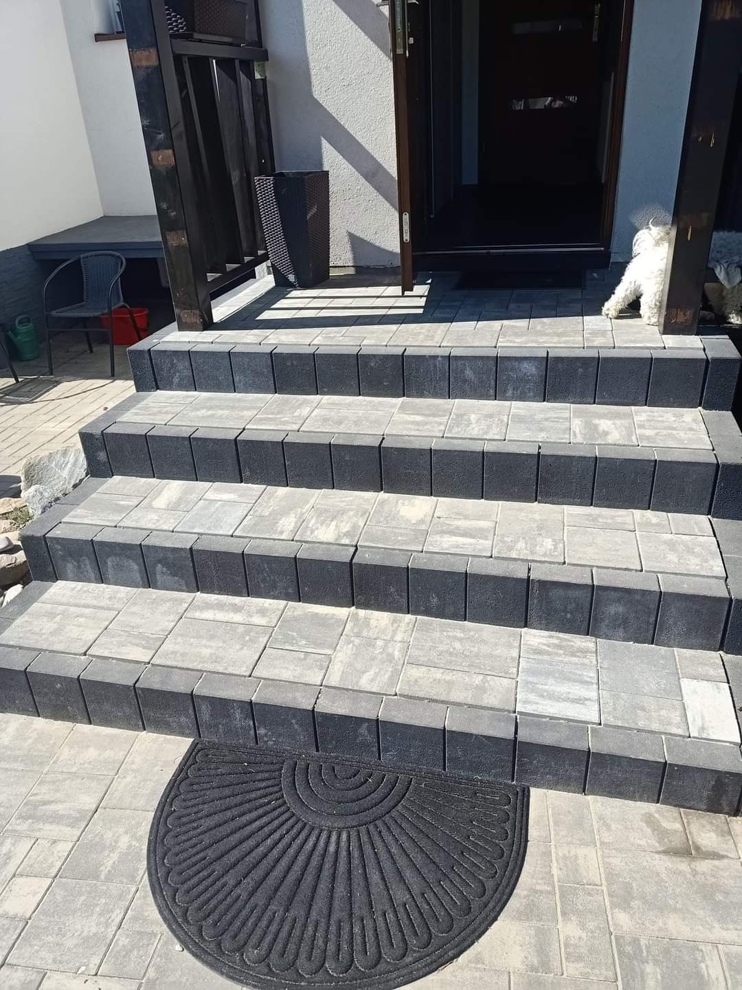Schody wejściowe wyłożone szarą kostką brukową z ciemnymi obrzeżami, z wycieraczką w kształcie wachlarza u dołu i białym psem w tle.