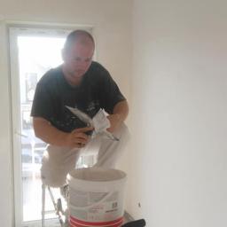 Domelektro - Mężczyzna szpachluje sufit w nowym mieszkaniu, stojąc na aluminiowej drabinie z wiadrem gładzi i szpachelkami