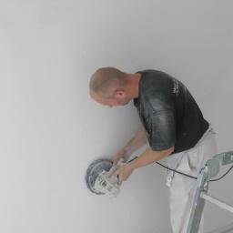 Domelektro - Mężczyzna szlifuje ścianę szlifierką elektryczną, stojąc na drabinie w pomieszczeniu w trakcie remontu, widoczne ślady pyłu na ubraniu.