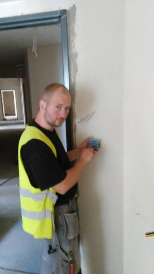 Mężczyzna w kamizelce odblaskowej szlifuje ścianę w nowym budynku, przygotowując ją do dalszych prac wykończeniowych.