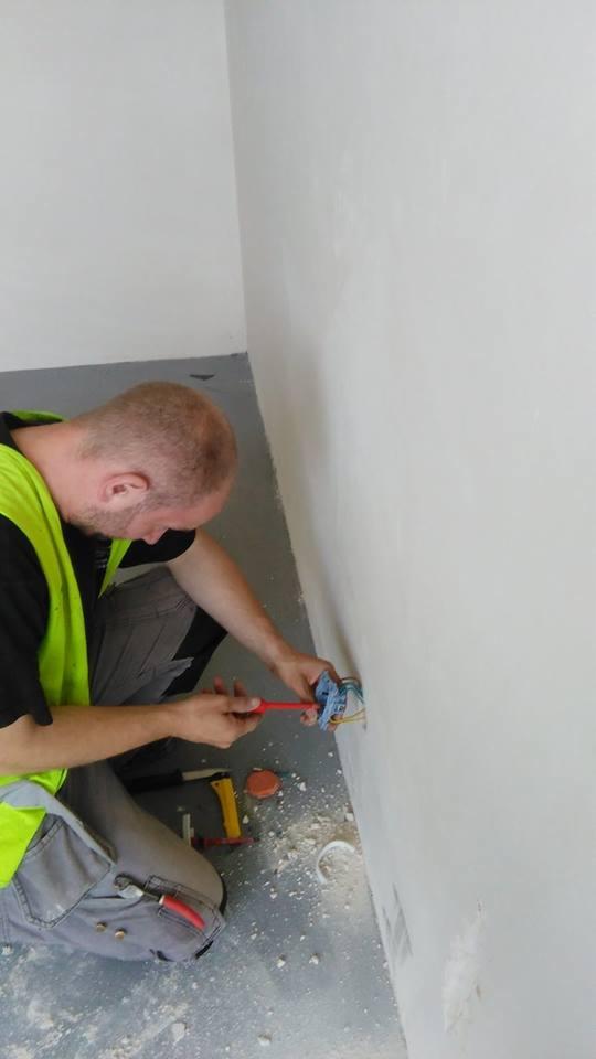 Elektryk w kamizelce odblaskowej, klęczący przy ścianie, używa śrubokręta do pracy przy przewodach elektrycznych wystających ze ściany, w pobliżu leżą narzędzia.