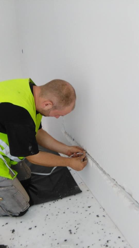 Mężczyzna w kamizelce odblaskowej szpachluje szczelinę między ścianą a podłogą, widoczne narzędzia i czarna mata ochronna na podłodze.