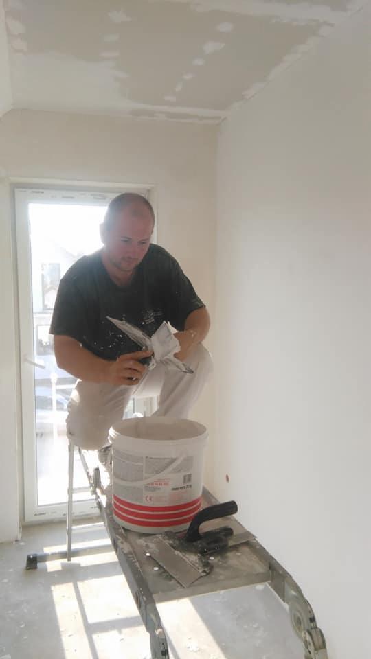 Mężczyzna szpachluje sufit w nowym mieszkaniu, stojąc na aluminiowej drabinie z wiadrem gładzi i szpachelkami