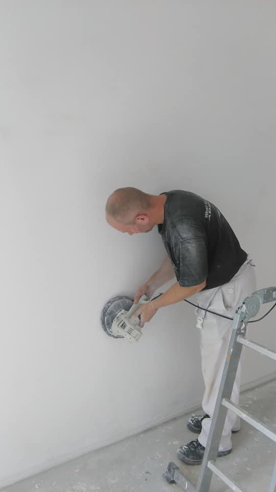Mężczyzna szlifuje ścianę szlifierką elektryczną, stojąc na drabinie w pomieszczeniu w trakcie remontu, widoczne ślady pyłu na ubraniu.