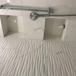 Biała łazienka z dekoracyjną, falistą płytką ścienną, chromowana bateria prysznicowa i wnęki wykończone płytkami imitującymi marmur.