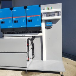 MIKRO3D-RAFAŁ HANKUS - Zestaw złożony z trzech niebieskich falowników Victron Energy MultiPlus-II, dwóch szarych akumulatorów oraz panelu elektrycznego z zabezpieczeniami, zamontowany na szarej platformie z regulowanymi...