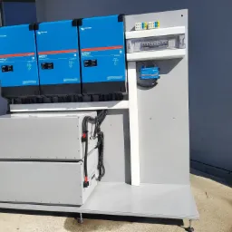 Zestaw złożony z trzech niebieskich falowników Victron Energy MultiPlus-II, dwóch szarych akumulatorów oraz panelu elektrycznego z zabezpieczeniami, zamontowany na szarej platformie z regulowanymi...