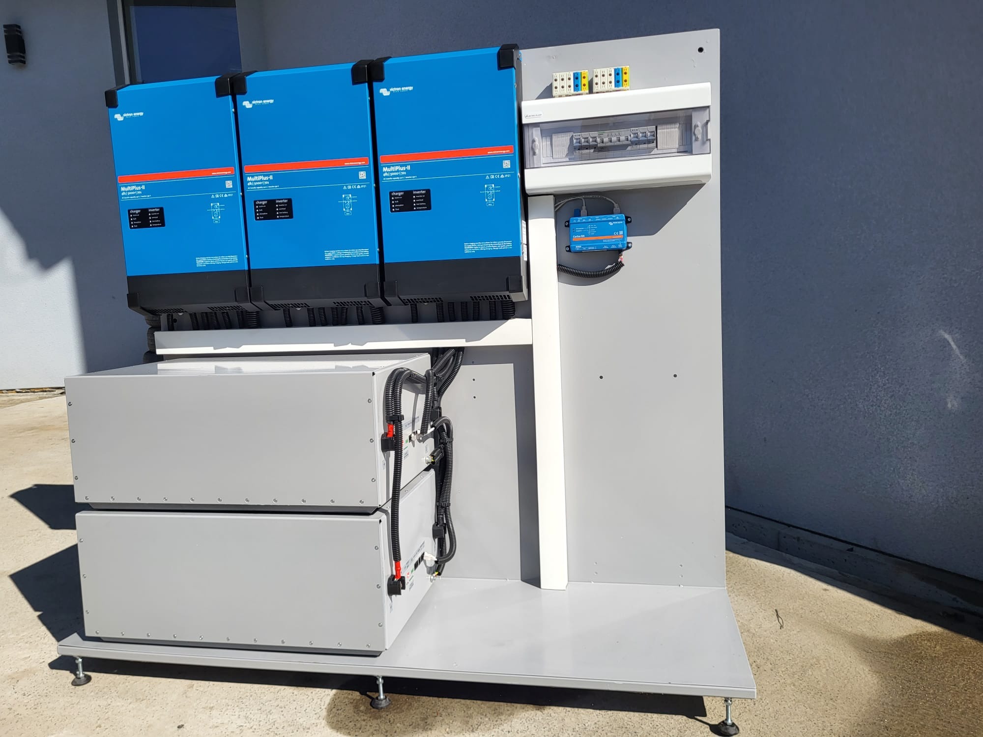 Zestaw złożony z trzech niebieskich falowników Victron Energy MultiPlus-II, dwóch szarych akumulatorów oraz panelu elektrycznego z zabezpieczeniami, zamontowany na szarej platformie z regulowanymi...