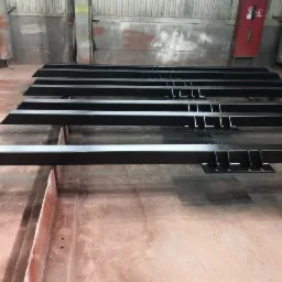 Czarne, stalowe belki z przyspawanymi elementami mocującymi, ułożone równolegle w hali produkcyjnej, gotowe do montażu.