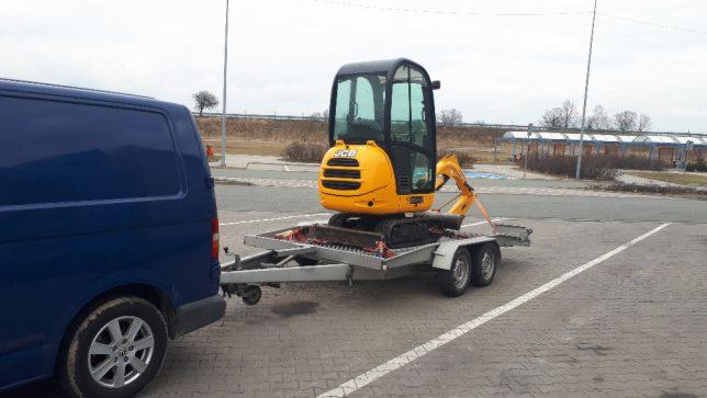 Żółta mini koparka JCB, zabezpieczona pasami, przewożona na przyczepie, podpiętej do niebieskiego vana na parkingu z widocznymi w tle wiatami przystankowymi.