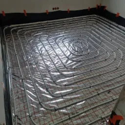 Białe rury ogrzewania podłogowego ułożone spiralnie na srebrnej folii z czerwoną kratką, przytwierdzone czarnymi klipsami, widoczne fragmenty ściany z izolacją przy podłodze.