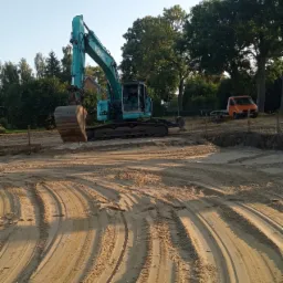 Turkusowa koparka Kobelco na piaszczystym terenie z widocznymi śladami opon, w tle drzewa i pomarańczowy samochód dostawczy.