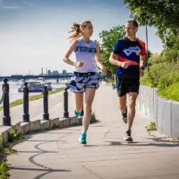 Kobieta i mężczyzna biegają obok rzeki w słoneczny dzień, widoczne budynki w tle, ona w koszulce Asics i wzorzystych spodenkach, on w kolorowej koszulce i czarnych spodenkach.