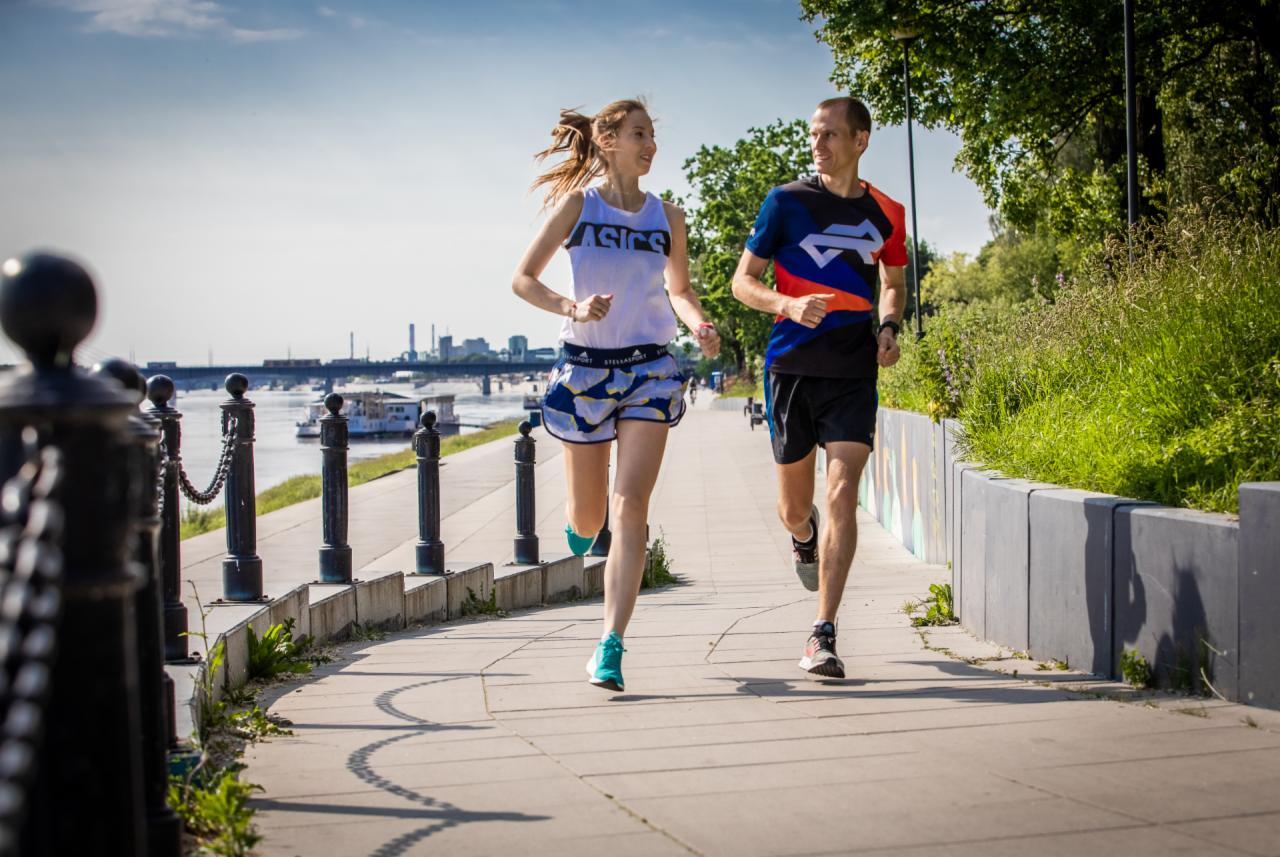 Kobieta i mężczyzna biegają obok rzeki w słoneczny dzień, widoczne budynki w tle, ona w koszulce Asics i wzorzystych spodenkach, on w kolorowej koszulce i czarnych spodenkach.