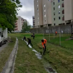Grupa robotników w odblaskowych kamizelkach i ciemnych ubraniach pogłębia rów melioracyjny na tle bloków mieszkalnych, z zaparkowanym busem w pobliżu.