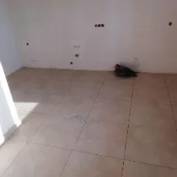 Świeżo ułożone, beżowe płytki podłogowe w pustym pomieszczeniu, widoczne niedoskonałości przy ścianach i mała poziomica na podłodze.