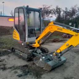 Żółto-szara mini-koparka Wacker Neuson na placu budowy o zmierzchu, z widocznymi śladami gąsienic i budynkiem w tle.