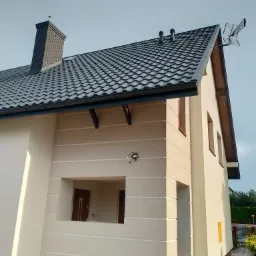 Jasna elewacja domu jednorodzinnego z ciemną, metalową dachówką, widoczne okna z brązowymi ramami, komin z cegły i antena satelitarna na dachu.