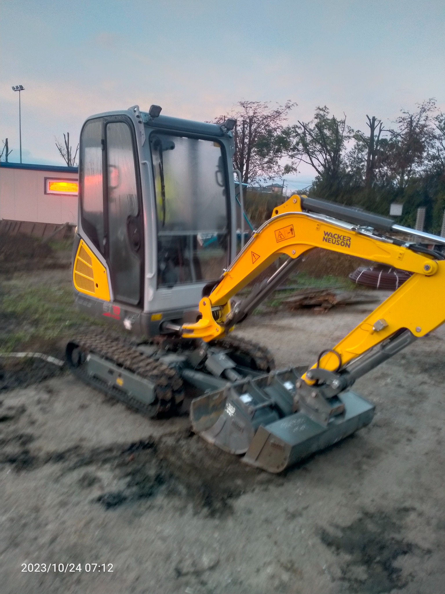 Żółto-szara mini-koparka Wacker Neuson na placu budowy o zmierzchu, z widocznymi śladami gąsienic i budynkiem w tle.