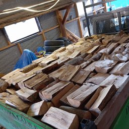 Drewno opałowe .Budowa Wiaty,Garaże z drewna ,Ogrodzenia panelowe oraz z drewna .Układanie kostki brukowej. - Kilkadziesiąt pociętych klocków drewna w przyczepie z zielonymi burtami, w tle opony, niebieska plandeka i konstrukcja dachu magazynu.