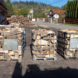 Drewno opałowe .Budowa Wiaty,Garaże z drewna ,Ogrodzenia panelowe oraz z drewna .Układanie kostki brukowej. - Trzy metalowe kontenery IBC wypełnione pociętym drewnem ułożone na żwirowym podłożu, w tle sterta drewna, zabudowania i zalesione wzgórza.