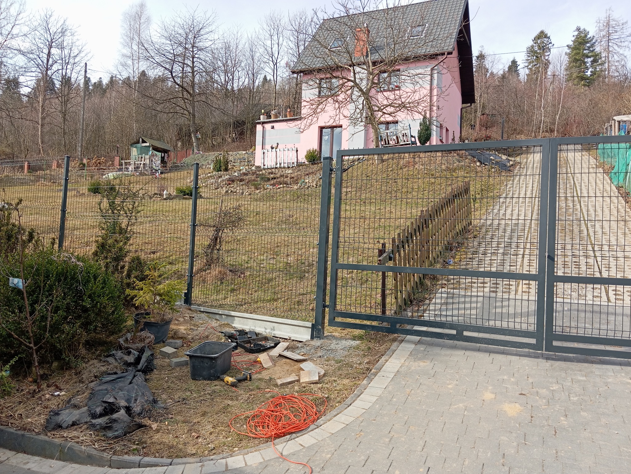 Montaż ogrodzenia panelowego i drewnianego w trakcie realizacji: widoczne narzędzia, kabel zasilający, czarna folia i sadzonki drzewek na tle domu z ogrodem.