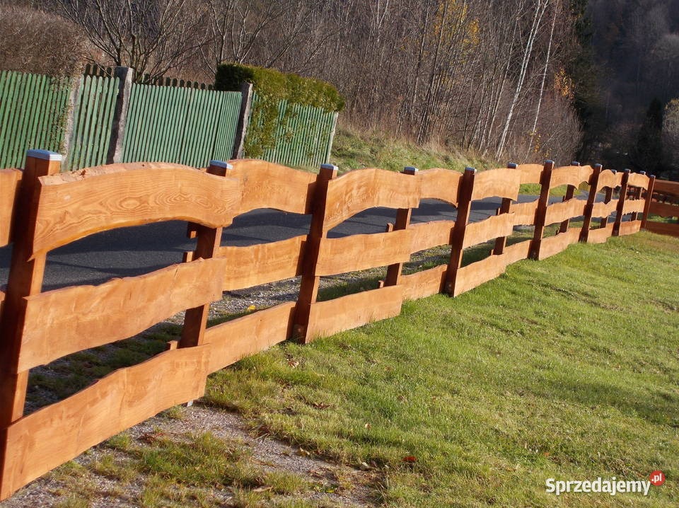 Dwuprzęsłowe, rustykalne ogrodzenie z surowego drewna w kolorze miodowym, oddzielające posesję od drogi asfaltowej, na tle zielonego trawnika i drzew.