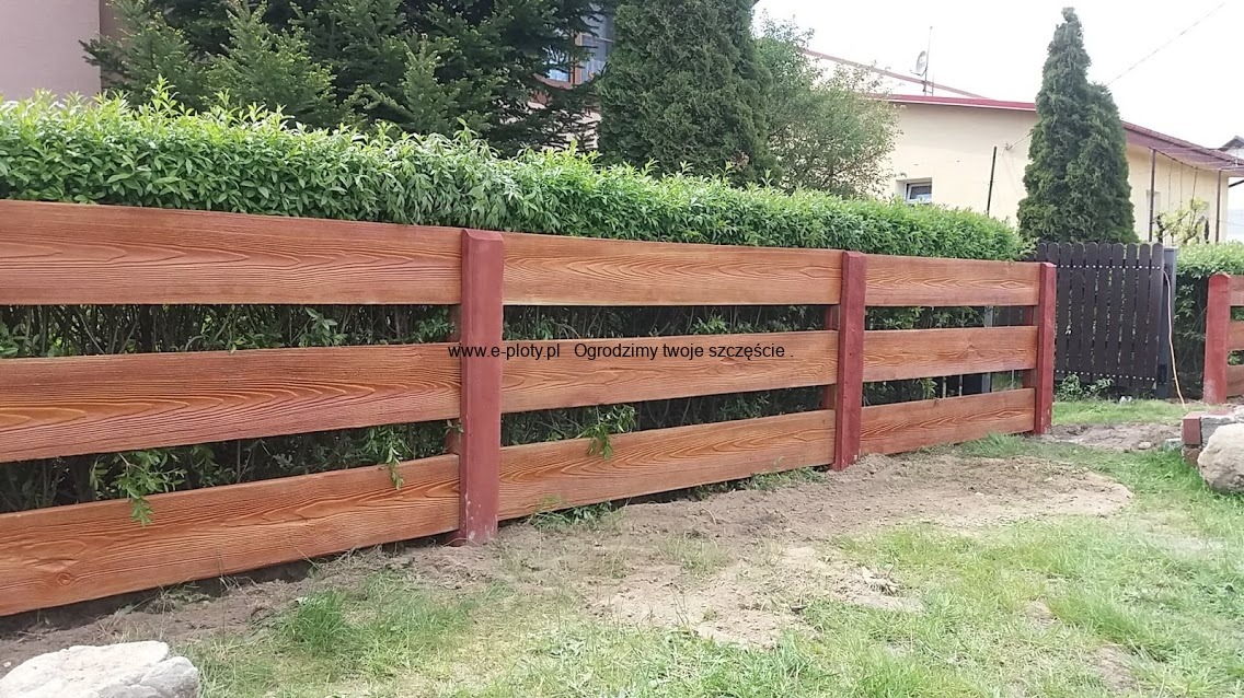 Drewniane ogrodzenie z poziomych desek o fakturze drewna i czerwonych słupkach, częściowo zasłonięte zielenią, z domem w tle.