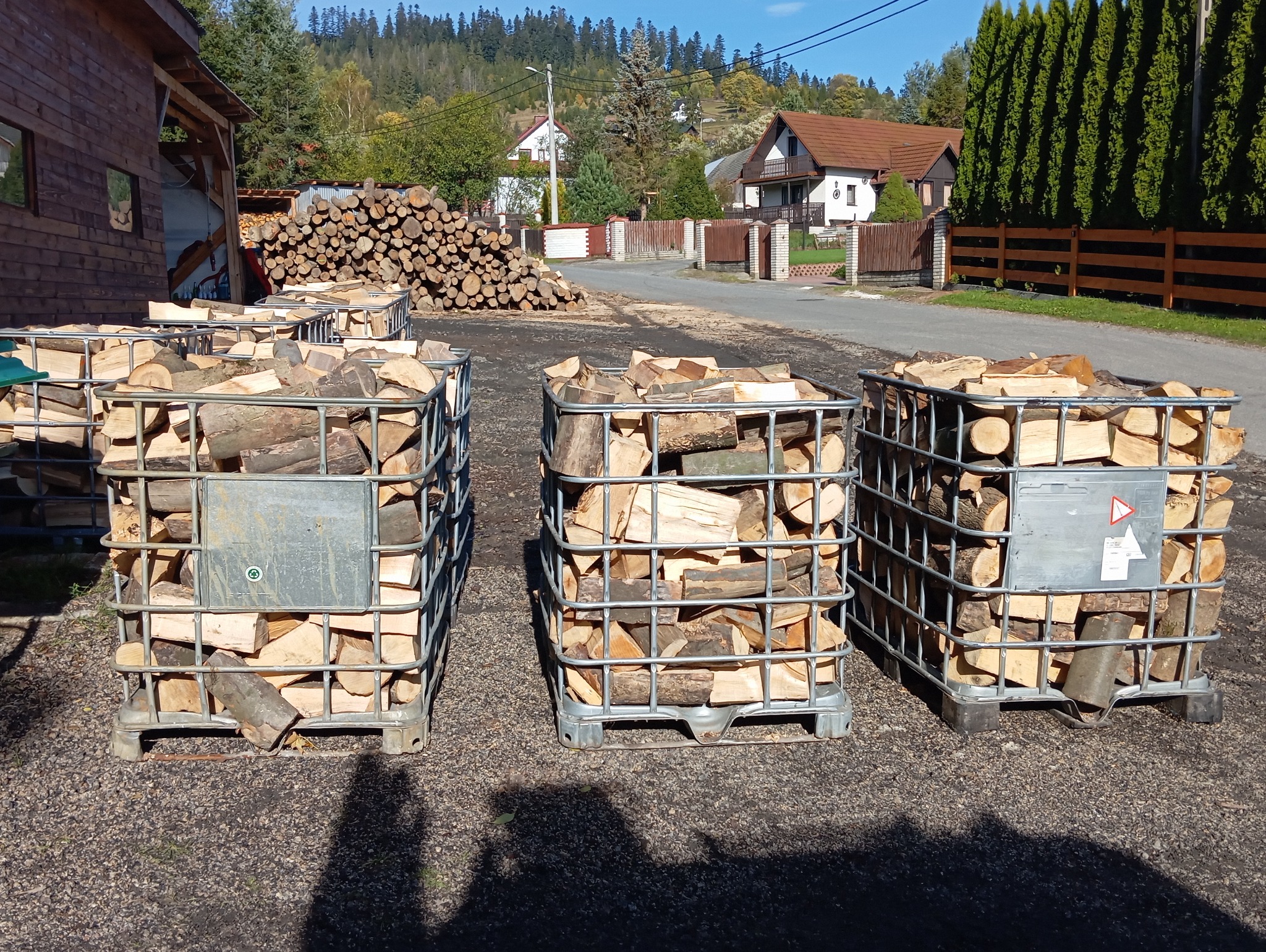 Trzy metalowe kontenery IBC wypełnione pociętym drewnem ułożone na żwirowym podłożu, w tle sterta drewna, zabudowania i zalesione wzgórza.