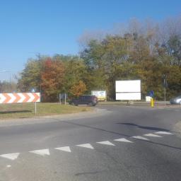 Berko - Oklejony billboard reklamowy przy rondzie z oznakowaniem poziomym i pionowym, w tle drzewa w jesiennej kolorystyce.