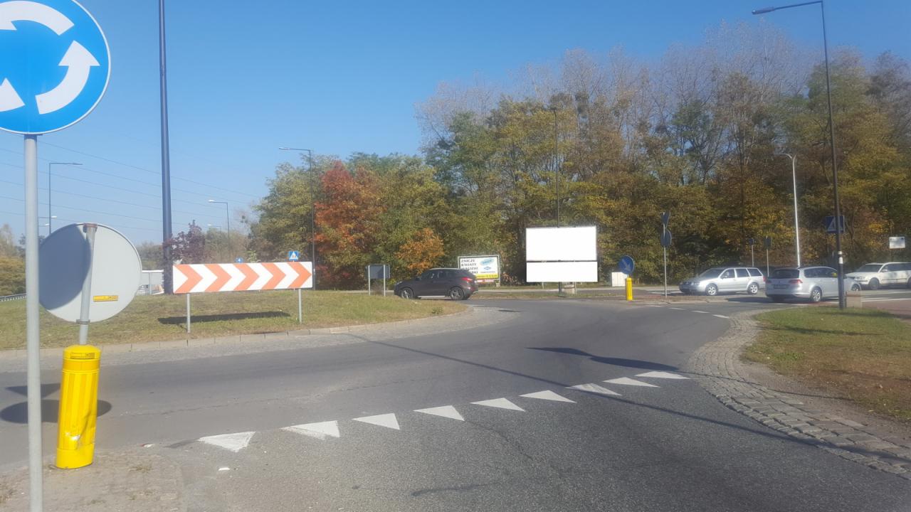 Oklejony billboard reklamowy przy rondzie z oznakowaniem poziomym i pionowym, w tle drzewa w jesiennej kolorystyce.