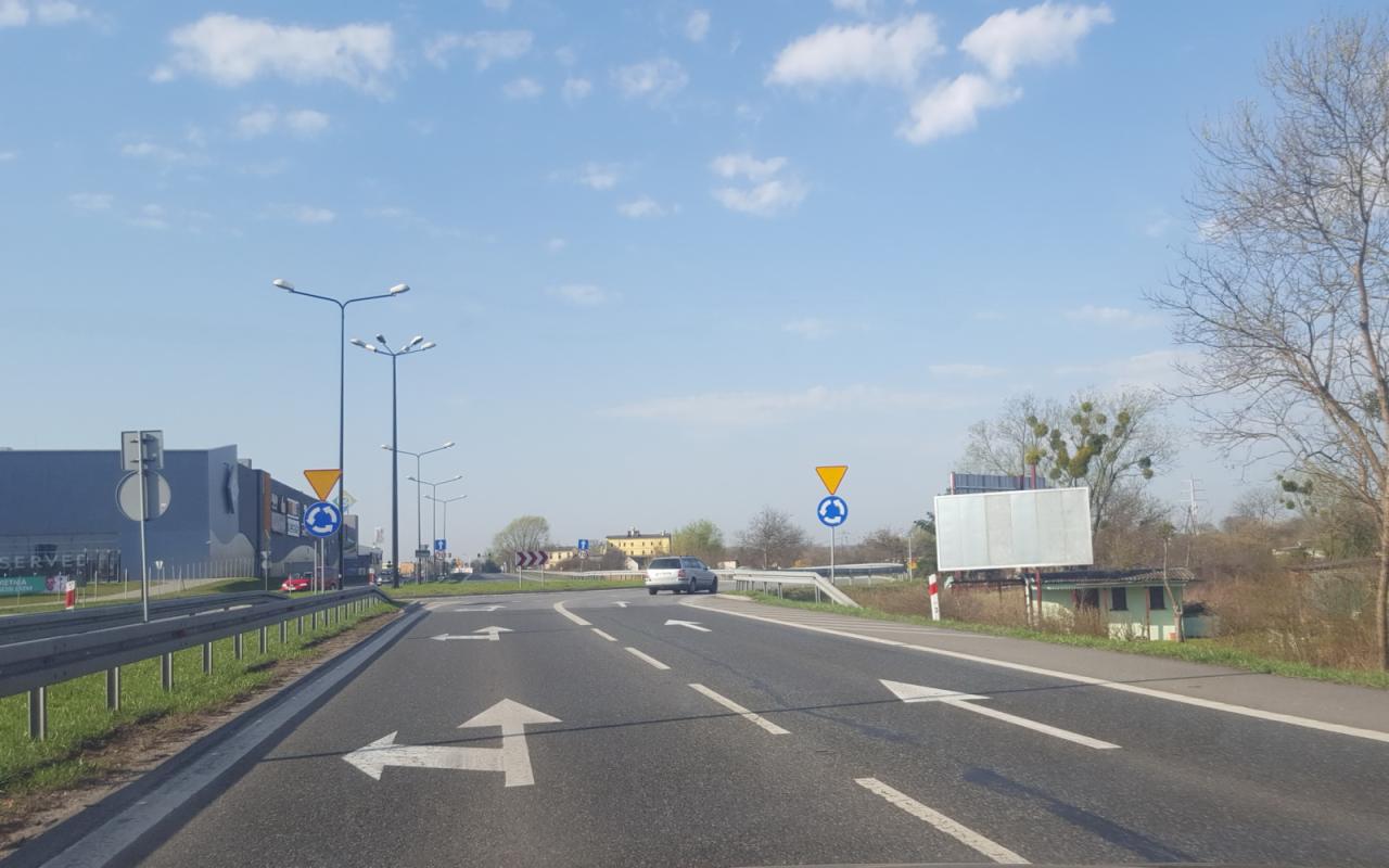 Droga z namalowanymi strzałkami kierunkowymi, znaki drogowe wskazujące rondo, budynek sklepowy w tle.