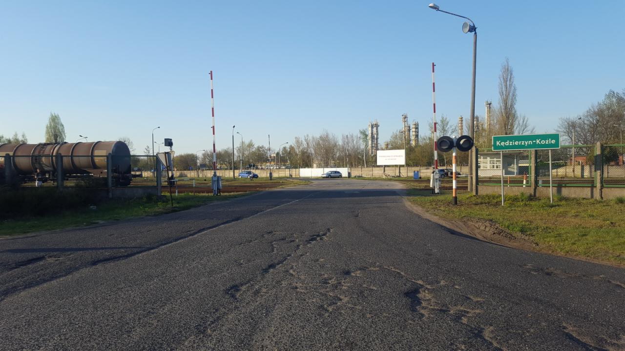 Przejazd kolejowy w Kędzierzynie-Koźlu z widocznymi cysternami kolejowymi, sygnalizacją świetlną i tablicą z nazwą miejscowości na tle zakładu przemysłowego.