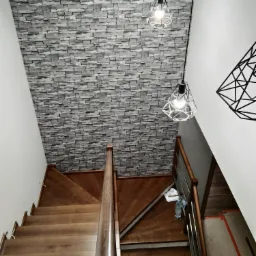 Drewniane schody z balustradą, ściana wykończona tapetą imitującą kamień, nowoczesne lampy wiszące z geometrycznym kloszem oraz dekoracja ścienna w kształcie rombu.